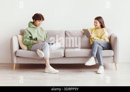 Handsome young Asian guy and his girlfriend using modern gadgets while sitting on couch at home, copy space Stock Photo