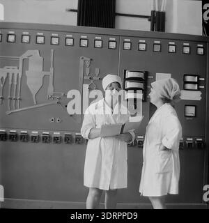 Historical 1960s black and white photograph captures two female technicians in white sanitary coats discussing production at the Slavyansk Oil and Fat Plant in the Ukrainian SSR, USSR. One worker holds a folder with documents while standing before a massive industrial control panel featuring gauges, switches, and a technical schematic diagram. This authentic archival image highlights Soviet engineering, the role of women in industry, and the optimistic atmosphere in the peaceful Donbass before the war, reflecting the region's economic development. Stock Photo