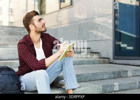 Young Man Writes in Notebook While Sitting on Steps of Modern Building During the Day Stock Photo