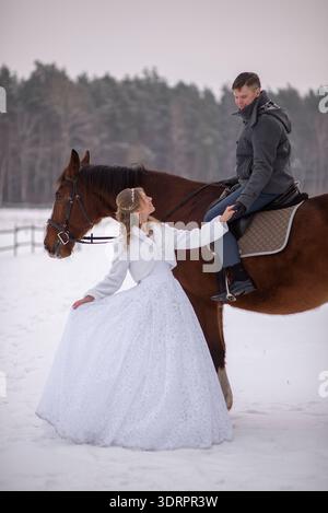 Bride stands beside a horse while the groom sits riding it, sharing a quiet and romantic winter wedding moment in a snowy rural landscape. Stock Photo