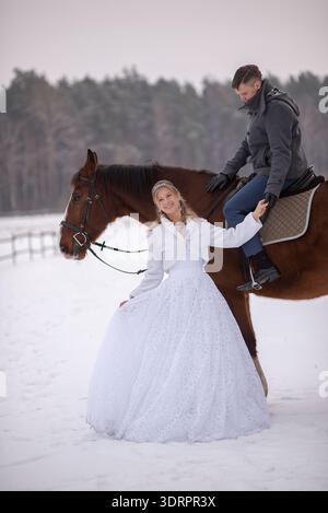 Bride stands still beside a brown horse while the groom sits on horseback behind her. She looks directly at the camera, creating a calm and elegant wi Stock Photo