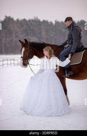 Bride stands beside a horse while the groom sits riding calmly. The scene captures a balanced and romantic winter wedding portrait in a snowy rural se Stock Photo