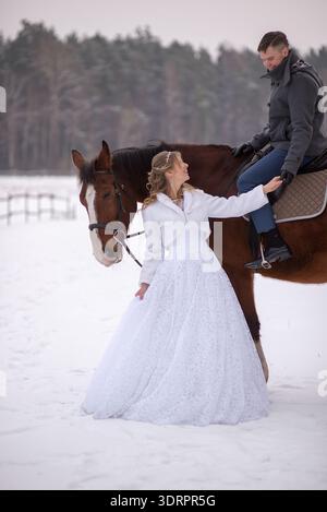 Bride in a white wedding dress stands beside a brown horse, gently reaching for the groom’s hand while he sits in the saddle during a winter wedding p Stock Photo