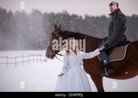 Bride stands beside a horse looking up at the groom while snow falls around them, capturing a romantic and atmospheric winter wedding scene. Stock Photo
