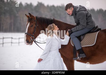 Smiling bride stands beside a horse, looking up at the groom with warmth and joy, creating a natural and romantic winter wedding moment. Stock Photo