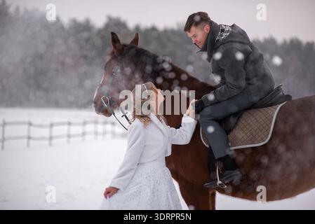 A bride in a white wedding dress stands beside a brown horse while the groom leans down from the saddle, holding her hand during a romantic winter mom Stock Photo