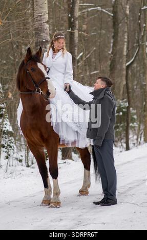 A groom stands beside a brown horse, carefully holding the brides hand as she prepares to dismount, creating a supportive and intimate winter wedding Stock Photo