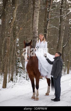Groom stands beside a brown horse while the bride sits on the saddle in a white wedding dress, smiling gently during a calm winter photoshoot in the f Stock Photo