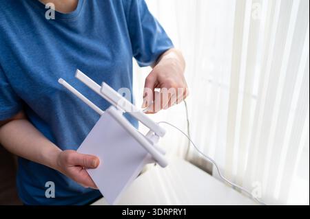 A woman in a blue shirt holds a wireless internet router while plugging in a network cable. This shot represents home network setup, technical install Stock Photo