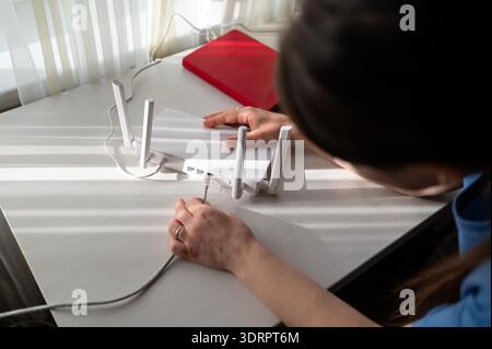 A young woman is setting up a wireless internet router with four antennas on a white wooden table near a window. Sunlight creates graphic shadows, rep Stock Photo