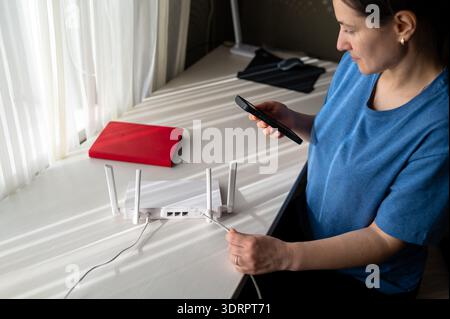 A person is holding a phone and setting up a wireless internet router with antennas on a white desk. This image represents smart home technology, mobi Stock Photo