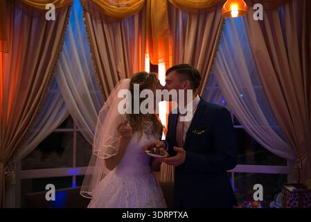 Bride and groom kissing while holding cake plates, warm evening lighting and soft interior colors creating intimate mood. Stock Photo