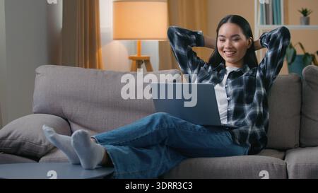Happy Asian Korean woman resting sofa watching movie laptop at home smiling freelance student female girl lying hands behind head work break enjoy Stock Photo