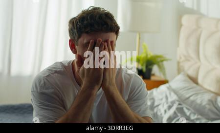 Despairing Caucasian man sitting on bed at home holding head in bedroom European male guy hopeless difficulties crisis grief anxiety distraught Stock Photo