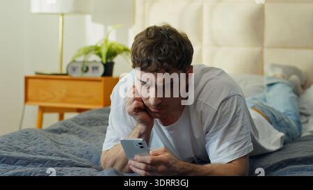 Worried Caucasian man using smartphone in bed at home frustrated male checking mobile phone problem error notification guy reading bad news banking Stock Photo