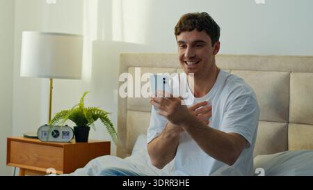Happy Caucasian man sitting bed in bedroom home talking smartphone recording video chat smiling male guy streaming online blog conference social Stock Photo