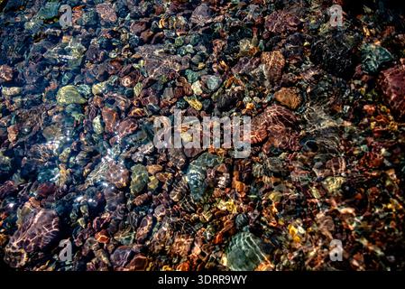 Transparent seawater flowing over smooth colorful pebbles along a Corsican shore, France, with shimmering light patterns, ideal for nature, travel, an Stock Photo