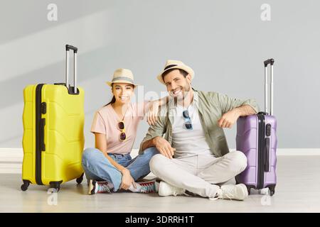 Smiling couple sitting on floor with suitcases in empty room, ready for summer trip and vacation. Stock Photo