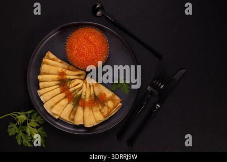 Delicious traditional blini crepes folded and topped with vibrant red caviar and fresh dill, served on a dark plate with a bowl of caviar and black cu Stock Photo