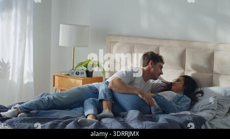 Caucasian man woman lying together on bed boyfriend girlfriend at home bedroom laughing smiling talking rest relaxing weekend comfortable pastime Stock Photo