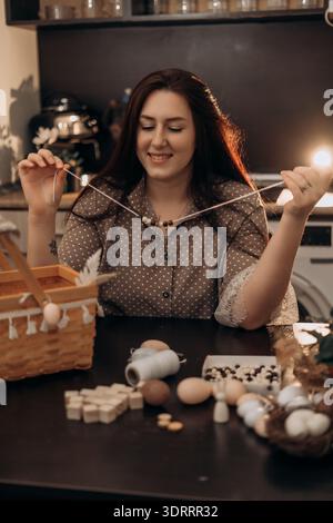 Woman stringing beads to make handmade necklace at home. Woman making handmade bead jewelry at kitchen table with craft supplies, smiling in warm Stock Photo