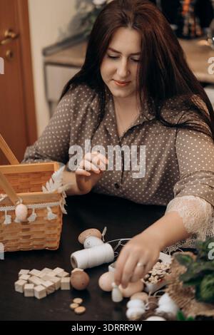 Woman crafting handmade holiday decor at home table. Woman at a table crafting holiday decorations with wooden beads, thread spools, and a wicker Stock Photo