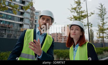 Contractors two engineers man woman talking looking at blueprint building plan diversity people colleagues builders architects wearing safety uniform Stock Photo