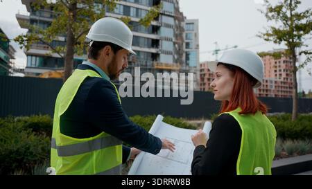 Two contractors civil engineers checking blueprint discuss buildings project teamwork woman man pointing city managers planning collaboration Stock Photo