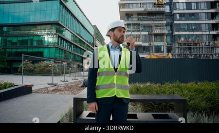 Moving shot Caucasian man builder engineer in hardhat talk remote radio outdoor urban building architect contractor wearing helmet safety vest Stock Photo