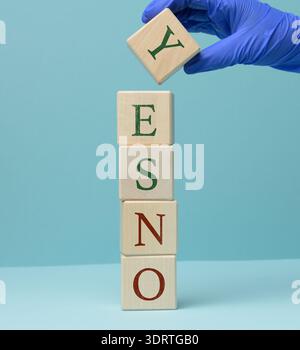 A hand in a blue medical glove places a wooden cube with the letter Y on a vertical stack of cubes, forming the word YES. Decision-making concept, med Stock Photo