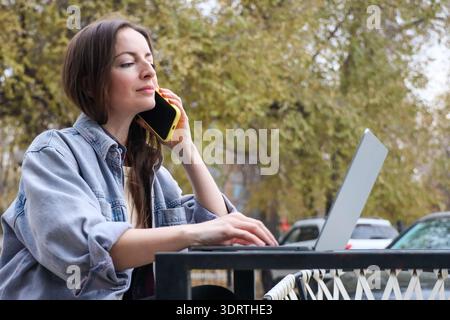 Mid adult woman working remotely on laptop while talking on smartphone at outdoor cafe terrace in autumn park Stock Photo