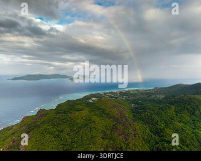 Aerial view of a vibrant rainbow arcs over the lush, verdant landscape and turquoise waters of Anse Reunion, La Digue, Seychelles. Stock Photo