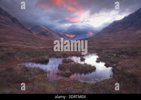 Dramatic sunset light over moorland pools in Glencoe, Scottish Highlands, with storm clouds and mountains in autumn. Stock Photo