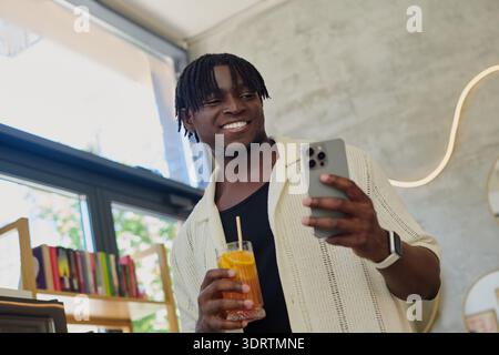 A young, handsome man holds a drink and smiles as he looks at his phone in a lively cafe. Stock Photo