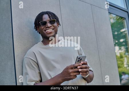 A cheerful young man with dreadlocks stands against a modern building wall, texting happily. Stock Photo