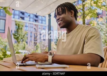 A young man is sitting outside, smiling as he uses his laptop under a canopy of trees. Stock Photo