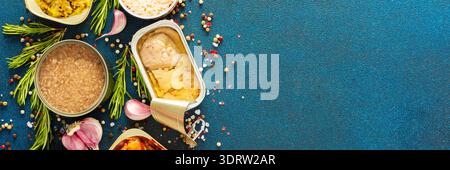 A top view of open tin cans with canned tuna, mackerel, shrimp, sardine, tuna, Cod liver, surrounded by spices and herbs on a blue background, banner Stock Photo