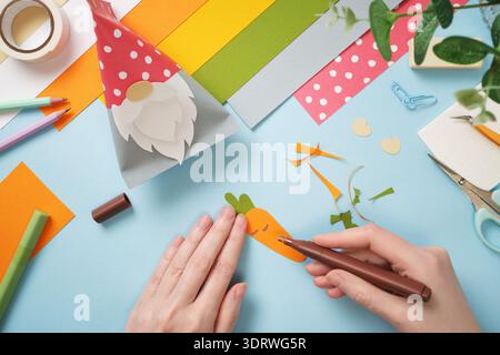Hands making paper gnome and carrot craft. Colorful paper, scissors, and markers on blue background. DIY handmade art project for kids. Stock Photo
