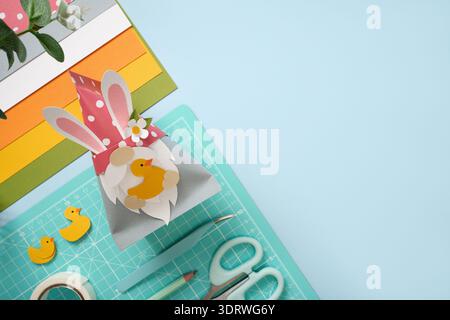 DIY Easter gnome with bunny ears and paper ducks. Scissors, cutting mat, and colorful paper on blue background. Copyspace Stock Photo
