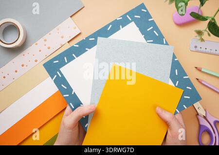 Hands holding colored paper for crafts. Tape, scissors, pencil on beige background. DIY handmade project for children. Stock Photo