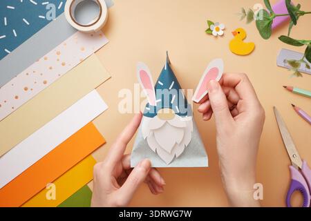 Hands attach rabbit ears to paper gnome. Tape, scissors, pencil on beige background. DIY handmade project for children. Stock Photo