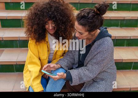 Two women of different backgrounds are seen seated on city steps, engrossed in their smartphones as they share content and bond through technology Stock Photo