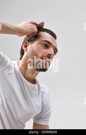 Low angle close up of man in his thirties with short beard combing hair with wooden comb on light gray background, Stock Photo