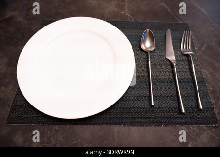 Empty white dinner plate with silverware on dark textured placemat, minimalist table setting Stock Photo