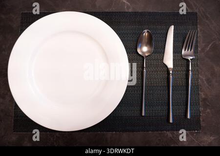 Empty white plate with silver cutlery on black textured placemat, top-down view, minimalist dining setup Stock Photo