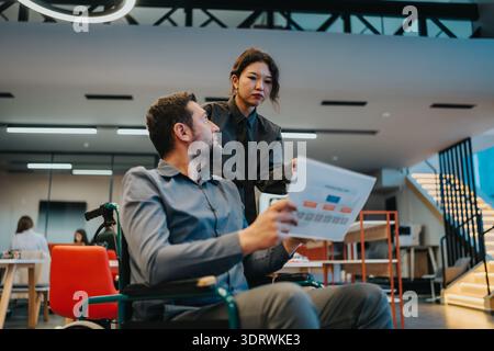Team collaboration in a modern office as a woman assists man in a wheelchair with documents Stock Photo
