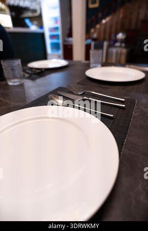 Empty white plates and silverware set on dark table in modern restaurant with blurred background Stock Photo