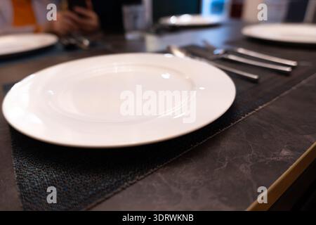 White ceramic plate on dark table with cutlery, empty restaurant setting, modern dining ambiance Stock Photo