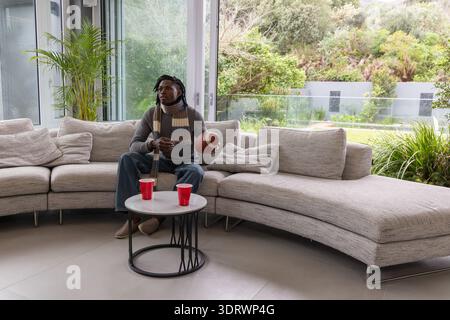 African American man sitting on gray sofa wearing striped scarf holding football checking phone Stock Photo