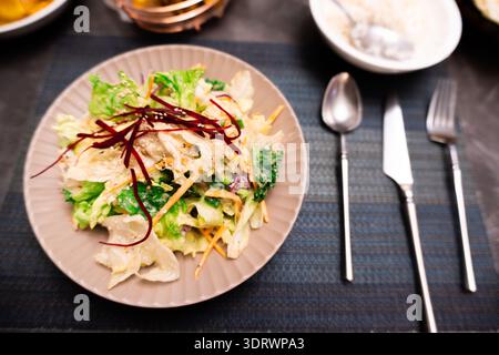 Asian-style salad with shredded vegetables and red garnish on ceramic plate, set on dark tablecloth with cutlery Stock Photo